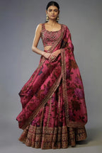 A model wearing a traditional Indian lehenga choli set in a rich magenta pink color with dark floral patterns and heavy gold embroidery, set against a plain gray studio background. A white label with 'D.