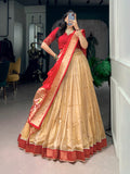 A woman wearing a traditional Indian lehenga choli outfit, featuring a beige pleated skirt with red borders, a red blouse with gold embroidery, and a red and gold dupatta, captured in a mirror selfie inside a boutique.