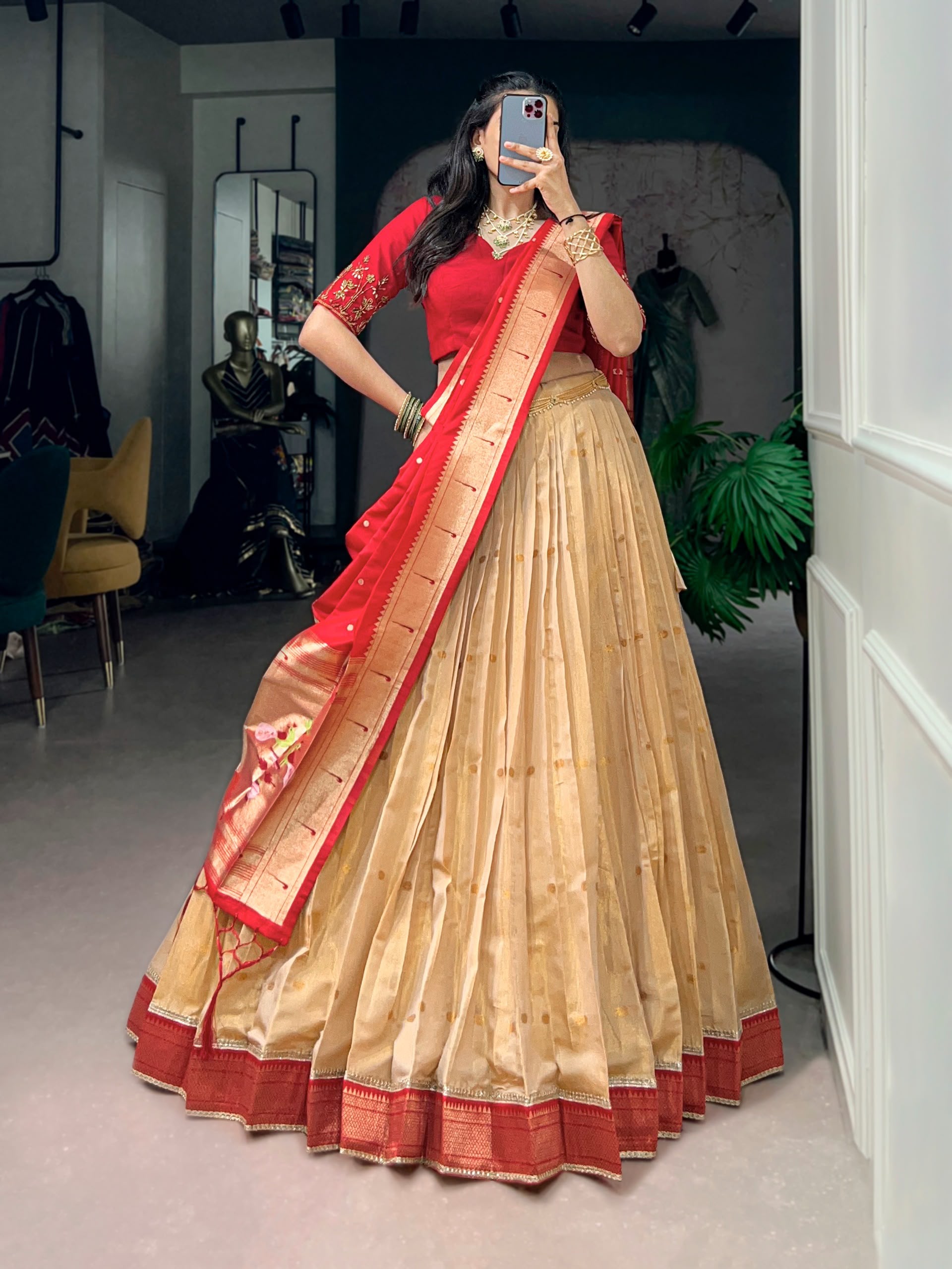 A woman wearing a traditional Indian lehenga choli outfit, featuring a beige pleated skirt with red borders, a red blouse with gold embroidery, and a red and gold dupatta, captured in a mirror selfie inside a boutique.