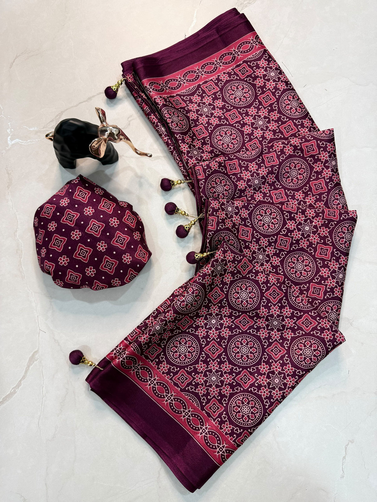 A folded traditional South Asian outfit set in deep magenta and purple, featuring a printed salwar fabric with circular mandala patterns, a matching small pouch, and a decorative black-and-gold shoe prop, laid flat on a light marble surface.