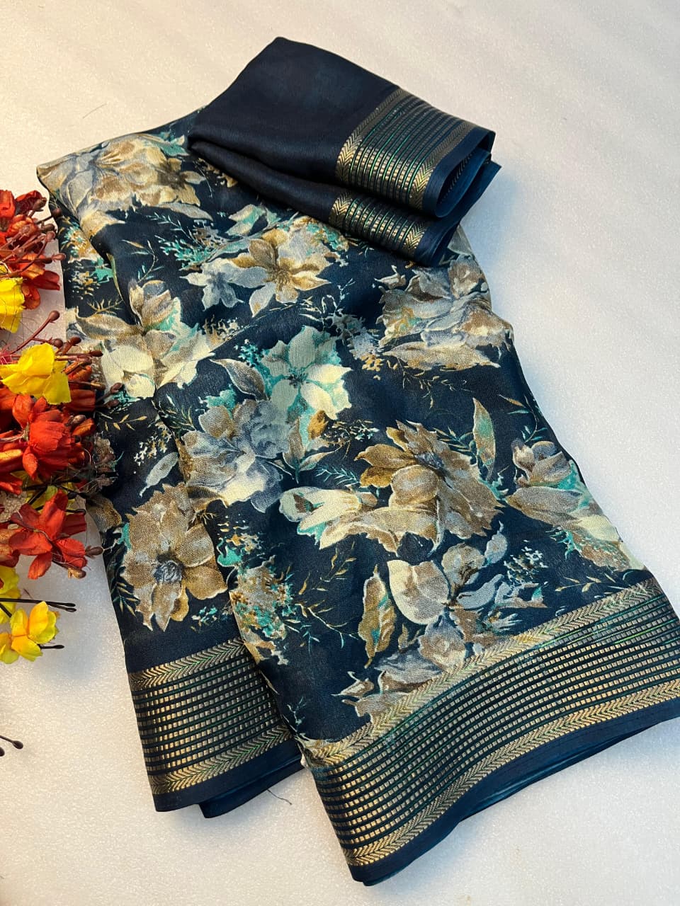A folded dark navy floral fabric, likely a saree or dupatta, with a gold zari border, displayed on a light beige surface next to decorative red and yellow flowers.