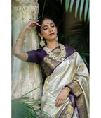 A woman wearing a traditional Indian saree leans against an ornate stone pillar in a garden setting. The saree has a metallic silver body with a deep purple border and gold zari embroidery, paired with a matching purple blouse and ornate gold jewelry.