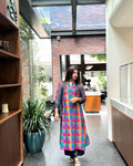A woman wearing a vibrant, multicolored plaid kurta with sheer long sleeves walks through a modern indoor hallway with a brick wall and glass skylight in the background.