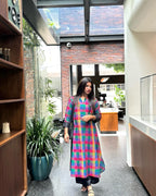 A woman wearing a vibrant, multicolored plaid kurta with sheer long sleeves walks through a modern indoor hallway with a brick wall and glass skylight in the background.
